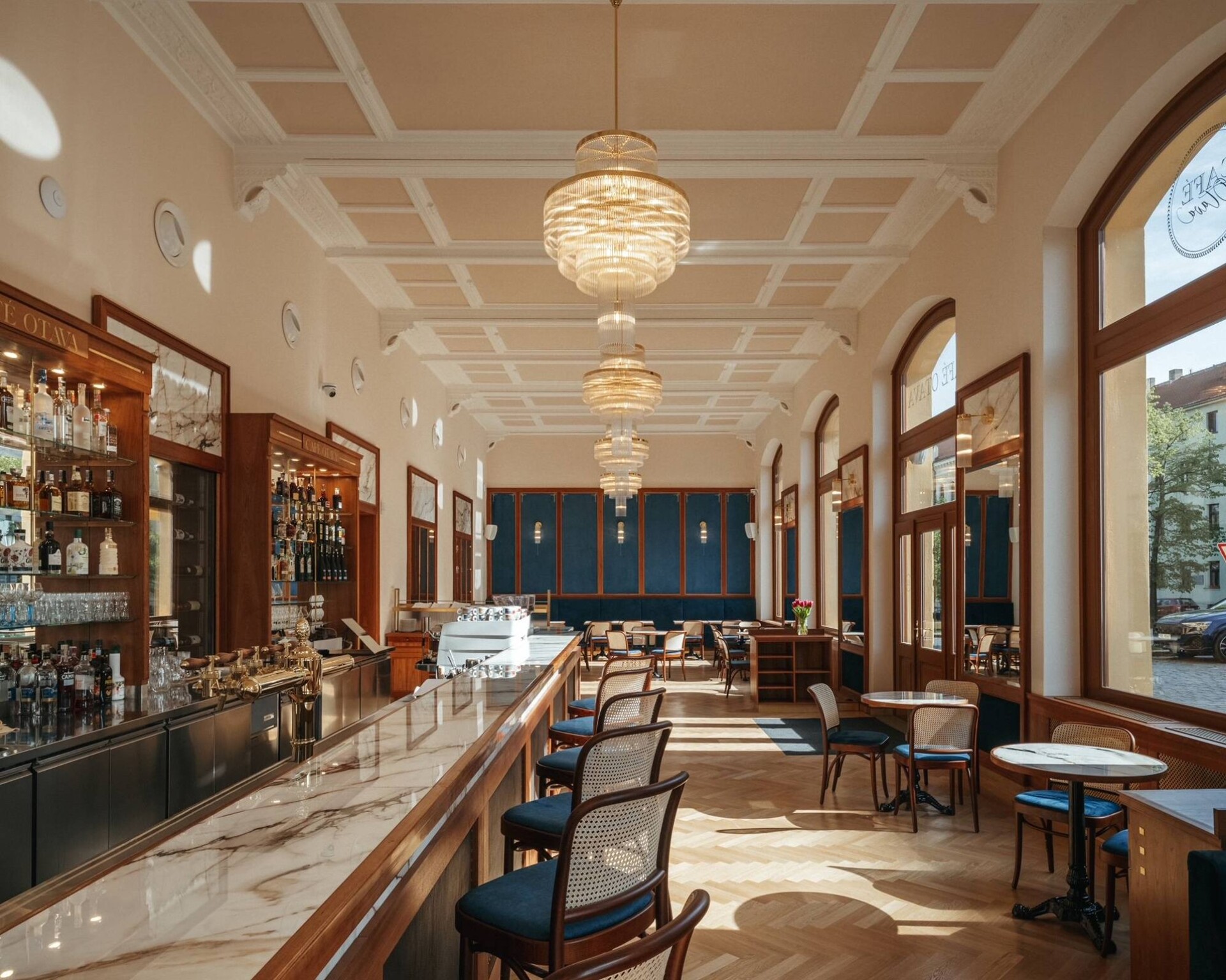 Entrance bar of Café Otava with a marble top, timber bar counter and three brass chandeliers beneath the coffered ceiling